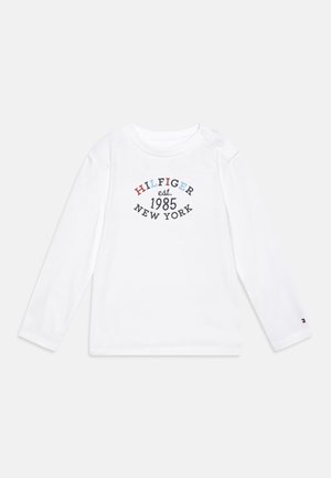 Camicia bianca a maniche lunghe con un design grafico colorato che riporta la scritta "HILFIGER est. 1985 NEW YORK" vicino al centro; dettaglio a scatto sulla spalla.