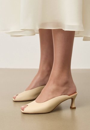 Nohy v béžových otvorených mule na podpätku v kombinácii s voľnou bielou midi sukňou, ktoré stoja na hladkej podlahe.