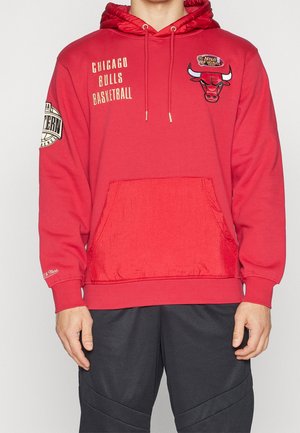 Czerwona bluza z kapturem Chicago Bulls z naszywką NBA Finals, logo drużyny na piersi oraz naszywką NBA Eastern Conference na lewym rękawie, noszona przez stojącą osobę.