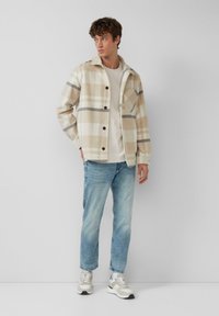 Beige en crème geruite shirtjack met donkergrijze accenten, knoopsluiting en borstzak; gecombineerd met lichtblauwe jeans en beige sneakers.