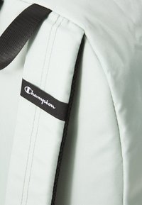 Sac à dos vert clair en tissu texturé ; pr�ésente des accents noirs et une étiquette de logo avec un texte blanc. Bandes noires ajustables incluses.