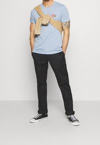 Světlomodré bavlněné tričko s grafickým potiskem, khaki mikina uvázaná na krku, černé kalhoty a černo-bílé tenisky.