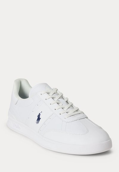 Witte lage sneaker met veters, stikseldetails en een klein donkerblauw Polo-logo aan de zijkant, afgebeeld tegen een effen achtergrond.