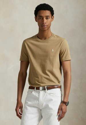 Giovane uomo che indossa una camicia beige a maniche corte con un piccolo logo e pantaloni bianchi con cintura marrone, in piedi davanti a uno sfondo uniforme.