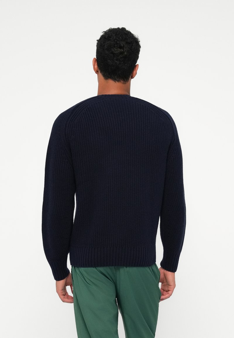 Marinblå ribbad stickad tröja med långa ärmar, rund halsringning och elastiska ärmslut; kombinerad med gröna byxor. Texturen verkar mjuk.