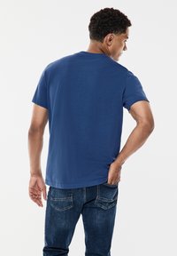 Sininen puuvillainen t-paita, jossa on pyöreä kaula-aukko ja lyhyet hihat, näytetään takaa. Yhdistetty tummansinisistä farkuista, joissa on klassinen malli ja takataskut.