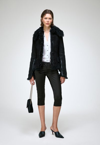 Femme portant une veste noire à col en fourrure, chemise blanche boutonnée, pantalon noir raccourci, talons noirs, tenant un petit sac à main noir avec chaîne dorée.