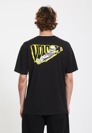 Persona con capelli ricci che indossa una maglietta nera con uno scheletro reclinato e la scritta gialla "VOLCOM" sul retro, su uno sfondo semplice.