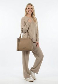 Lächelnde Frau in beigefarbenem Pullover und Hose hält eine taupefarbene Handtasche über der Schulter und trägt weiße Sneaker vor einem einfachen weißen Hintergrund.