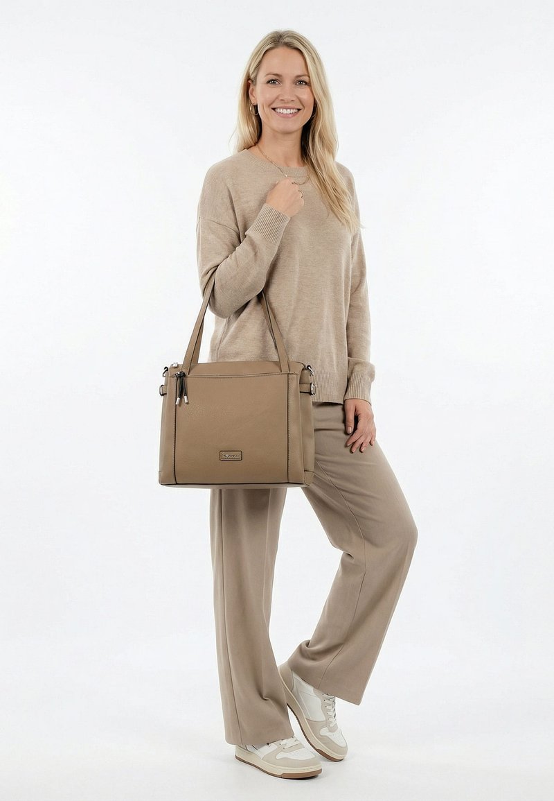 Lächelnde Frau in beigefarbenem Pullover und Hose hält eine taupefarbene Handtasche über der Schulter und trägt weiße Sneaker vor einem einfachen weißen Hintergrund.