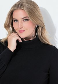 Černý rolák se žebrovanými manžetami, modelka s dlouhými vlnitými blond vlasy, nosí velké stříbrné kruhové náušnice, jemně se usmívá.