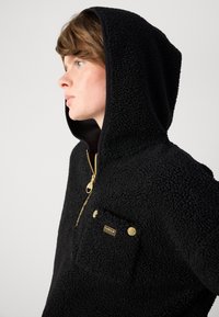 Mala de capuz em fleece preto com uma superfície texturizada, fecho na frente, detalhes em dourado e um bolso no peito com o logotipo da marca.
