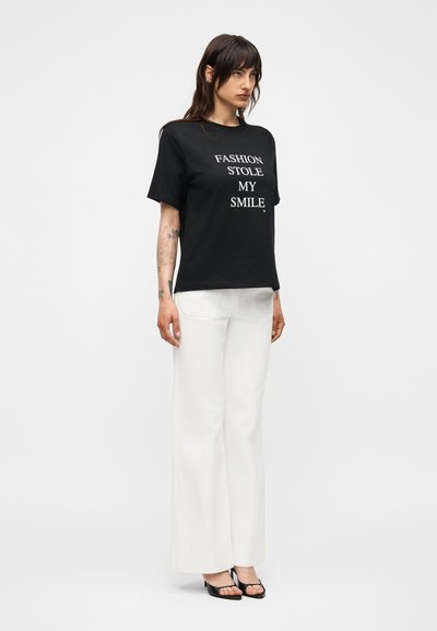 Γυναίκα με μακριά σκούρα μαλλιά φορώντας μαύρο T-shirt με το κείμενο «FASHION STOLE MY SMILE», άσπρο παντελόνι με φαρδιά πόδια και μαύρες ανοιχτές γόβες, όρθια.