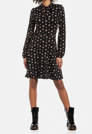 Femme portant une robe noire à manches longues à motifs floraux avec un col noué à l'avant, assortie à des bottines de combat noires brillantes à lacets.