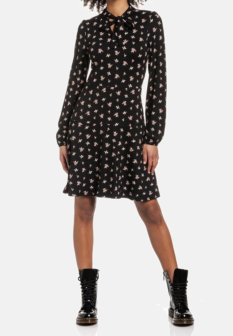 Femme portant une robe noire à manches longues à motifs floraux avec un col noué à l'avant, assortie à des bottines de combat noires brillantes à lacets.