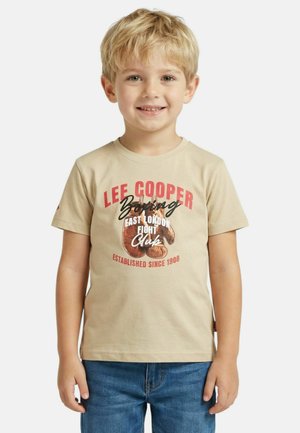 Jeune garçon souriant, portant un T-shirt beige à thème de boxe Lee Cooper et un jean bleu, debout devant un fond blanc.