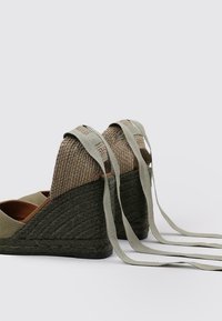 Espadrilles à talon compensé en tissu vert olive, avec un talon en chanvre tissé et de longues lanières assorties. Dessus en toile texturée, semelle en caoutchouc robuste et couture détaillée.