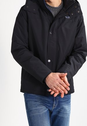 Mand iført en sort jakke over en grå sweater og blå jeans, med hænderne foldet foran sig mod en hvid baggrund.