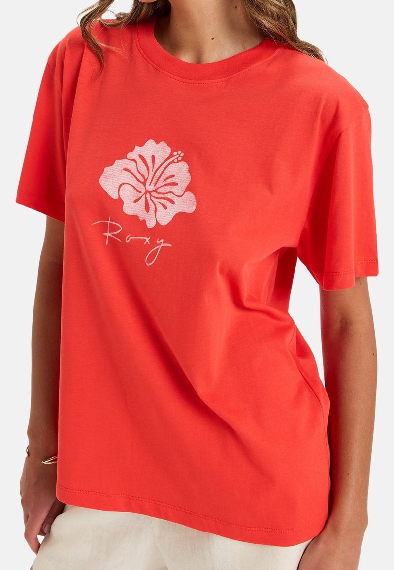 Femme portant un T-shirt rouge vif à manches courtes avec une fleur d'hibiscus blanche et le texte "Roxy", associé à un pantalon clair.