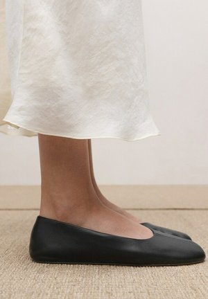 Pies con zapatos planos negros de punta cuadrada, con una falda midi color crema, sobre una alfombra beige y un fondo de pared clara.