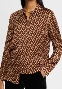 Blouse col chemise en tissu soyeux, présentant un motif géométrique bordeaux, jaune et noir sur fond beige. Manches longues, fermeture à boutons.