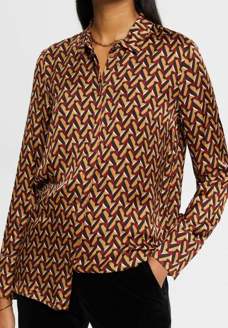 Blouse col chemise en tissu soyeux, présentant un motif géométrique bordeaux, jaune et noir sur fond beige. Manches longues, fermeture à boutons.