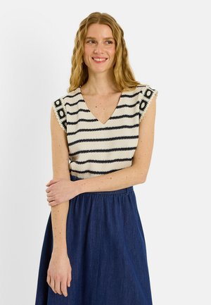 Femme aux cheveux blonds ondulés portant un haut en crochet rayé noir et blanc sans manches et une jupe bleue taille haute, souriante et regardant sur le côté.