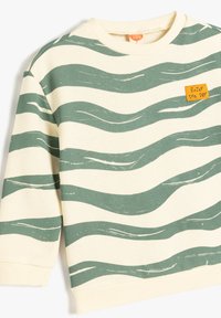 Sudadera beige claro con patrón de olas verdes. Cuenta con una etiqueta amarilla que dice "Disfruta el día." Textura suave y puños acanalados.
