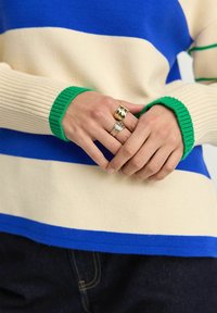 Gebreide trui met horizontale strepen in blauw, crème en groen. De mouwen hebben een geribbelde textuur, versierd met ringen op de handen.