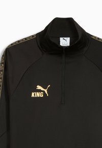Svart zip-up sportjacka i mjukt material, med gyllene detaljer, tryck med "KING" och PUMA-logotyp. Geometriskt mönster på axlarna.