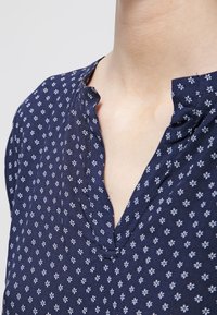 Blusa in tessuto blu navy con placca frontale, caratterizzata da una stampa floreale bianca. Texture morbida con vestibilità rilassata e maniche corte.