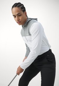 Sweat-shirt léger de sport avec des panneaux de couleur grise et blanche, texture lisse, fermeture éclair complète et capuche avec cordon de serrage, associé à un pantalon noir ajusté.