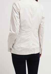 Giacca aderente color beige chiaro con una texture liscia, dettagli di cucitura sottili e polsini con bottoni. Presenta un colletto alto e una silhouette su misura.