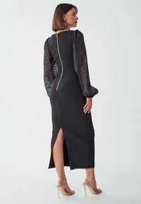 Čierne midi šaty s priesvitnými rukávmi s bodkami, so zipsom na zadnej strane a bočným rozparkom. Elegantný dizajn s priliehavým strihom a hladkou textúrou.