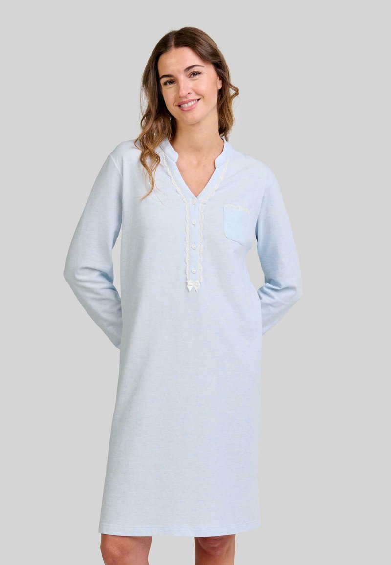 Mujer sonriendo con cabello ondulado que lleva un vestido de noche de manga larga y hasta la rodilla de color azul claro, con botones y detalles de encaje.