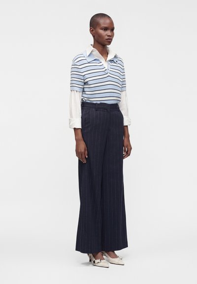 Femme portant un pull à manches courtes rayé bleu clair et blanc sur une chemise blanche à col, avec un pantalon large à fines rayures bleu marine et des talons blancs.