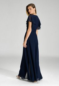 Abito blu navy con maniche a volants, corpetto arricciato e gonna lunga, caratterizzato da una sovrapposizione trasparente e scarpe con tacco argento minimaliste.
