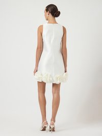 Γυναίκα με αμάνικο άσπρο μίνι φόρεμα με μεγάλες λεπτομέρειες από ύφασμα λουλουδιών κατά μήκος του τελειώματος, φορώντας λευκά ψηλοτάκουνα παπούτσια, με την πλάτη στραμμένη.