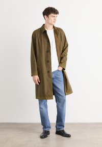 Olive green button-up coat with large buttons, two side pockets, and a relaxed fit, paired with a white shirt and blue jeans.
