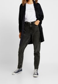 Zwarte denim hoge taille jeans, relaxte pasvorm; gecombineerd met een witte t-shirt met een zwart lijntekening. Donkere, oversized jas en blauwe sneakers.
