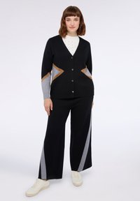 Zwarte cardigan met grijs en bruin zigzagaccenten, knoopsluiting, gecombineerd met brede zwarte broek met grijze zijstrepen en witte sneakers.