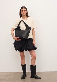 Donna che indossa una giacca corta a maniche corte colore crema, una gonna a palloncino nera, stivali neri al ginocchio, mentre tiene una grande borsa in pelle nera contro un muro semplice.