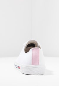 Weißer Sneaker mit niedrigem Schaft, pinkem Fersenlaschen und dunkelblauen sowie roten Streifen an der Sohle, von hinten auf weißem Hintergrund dargestellt.