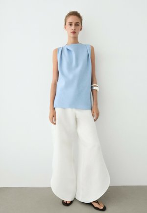 Donna con top azzurro chiaro senza maniche, pantaloni bianchi larghi, sandali neri e braccialetti bianchi, in piedi davanti a una parete chiara e semplice.