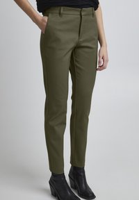 Olijfgroene broek met een gladde textuur, slim fit, riemlussen en zijzakken, gecombineerd met zwarte, hoge enkellaarsjes.