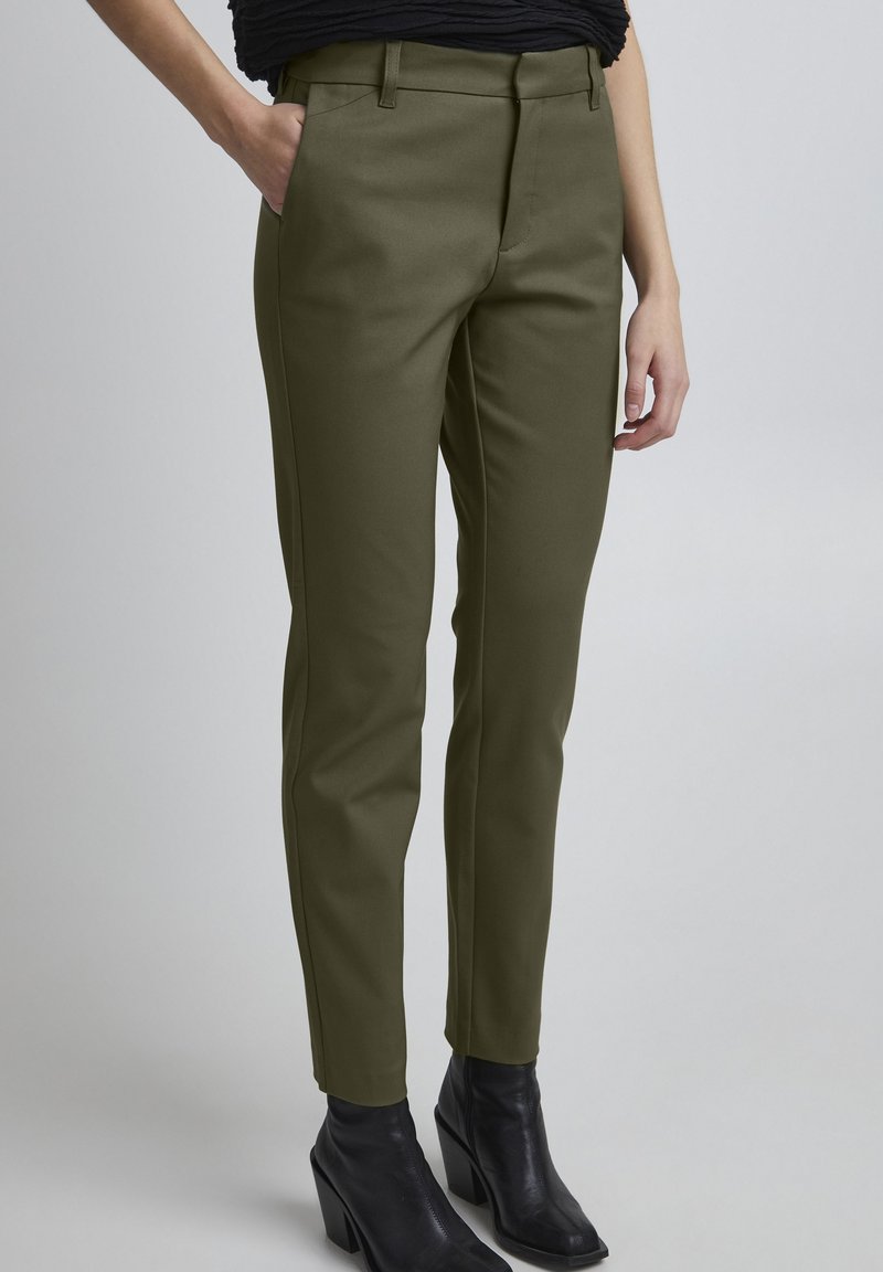 Olijfgroene broek met een gladde textuur, slim fit, riemlussen en zijzakken, gecombineerd met zwarte, hoge enkellaarsjes.