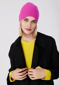Gorro de punto acanalado de color rosa brillante, llevado con un abrigo negro y un jersey amarillo. El gorro tiene una parte superior redondeada y un ajuste ceñido.