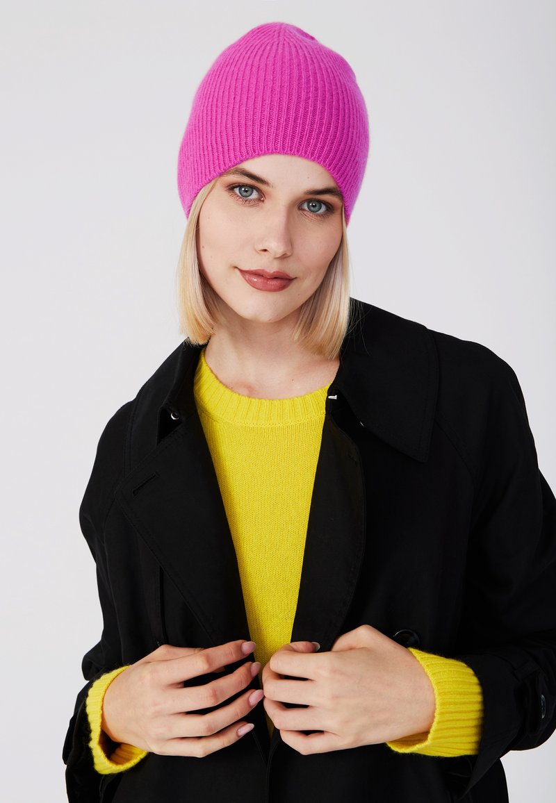 Gorro de punto acanalado de color rosa brillante, llevado con un abrigo negro y un jersey amarillo. El gorro tiene una parte superior redondeada y un ajuste ceñido.