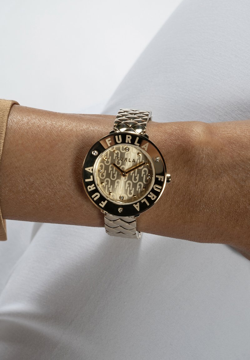 Orologio dorato con quadrante rotondo, dial a motivo e bracciale in metallo argentato con design intrecciato. Nome del marchio "Furla" in evidenza.