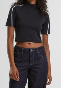 Svart kortärmad crop top med hög halsringning och vita sidostrikar, kombinerad med mörkblå jeans med kontrastsömmar och knappstängning.